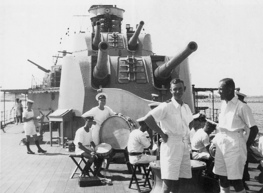 A view looking forward of Sydney's two aft 6-inch guns. In the foreground stands Lieutenant Commander R.D. Handcock (left) one of Sydney's engineering officers. Behind him members of Sydney's band can be seen practising.