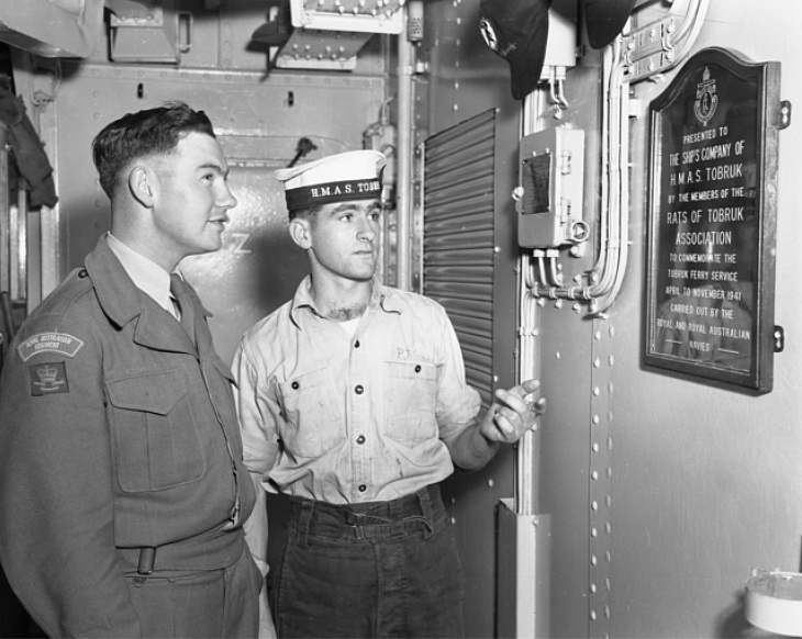 Private (Pte) Edwin Henry Cowley, 1st Battalion The Royal Australian Regiment (1RAR), of Murray Bridge, SA, is listening with interest to the history of the mess hall plaque. Able Seaman (AB) Peter Stokes, of Black Forest, Adelaide, SA, explains that all the crew are honorary members of the Rats of Tobruk Association which had adopted the ship in memory of the Tobruk ferry service in 1941