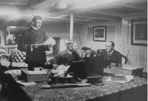 Assistant Paymaster Alfred Treacy (standing) onboard HMVS Nelson in 1897. The Captains Cabin, in which he is working, must have been a spacious compartment as Treacy was 6 feet and 1 inch tall and his head is well clear of the deckhead